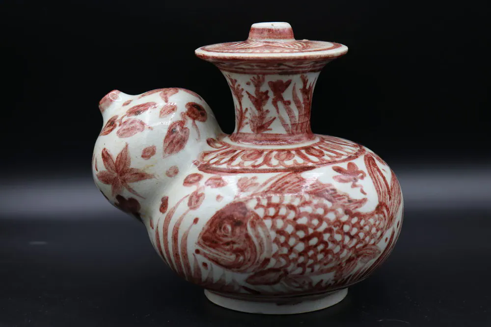 Three-quarter view of the copper-red kendi with a fish motif on the body and a floral spray on the domed spout