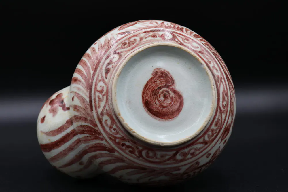 Base of the kendi showing a red-painted spiral shell mark within a scrolling border, spout knob at left