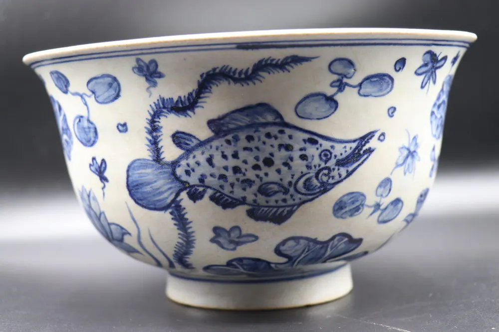 Front view of a blue and white bowl with a fish among lotus, ferns, and lily pads