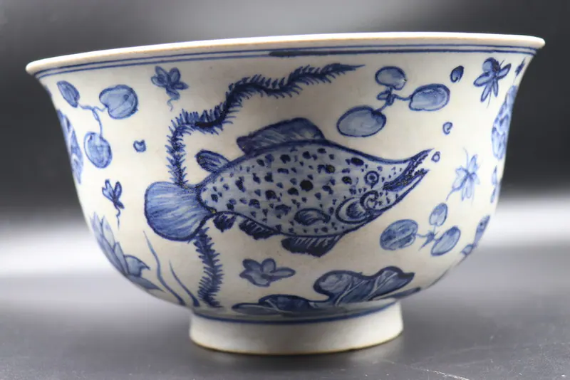 Front view of a blue and white bowl with a fish among lotus, ferns, and lily pads