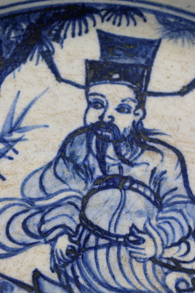 Close-up of a robed figure wearing a tall hat, holding a round drum, in underglaze blue