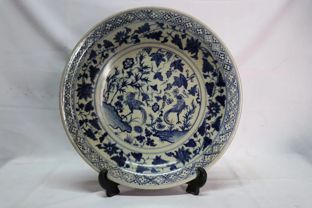 Front view of a blue and white dish on a stand, with two birds among rocks, vines and flowers