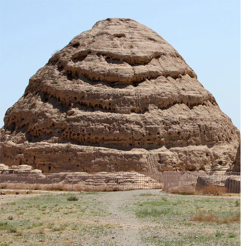 Large weathered earthen burial mound with eroded, layered sides behind a low fence