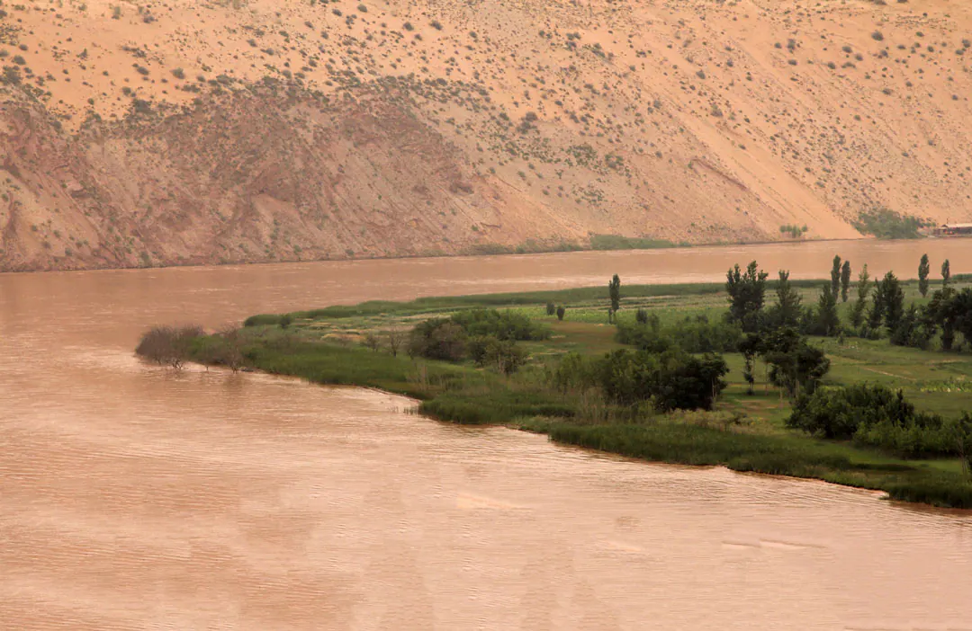 Silt-brown river curving past a green, tree-lined bank below a bare sandy hillside