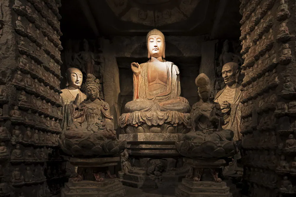 Painted stone Buddha seated between attendant figures in a cave shrine lined with small carved Buddhas