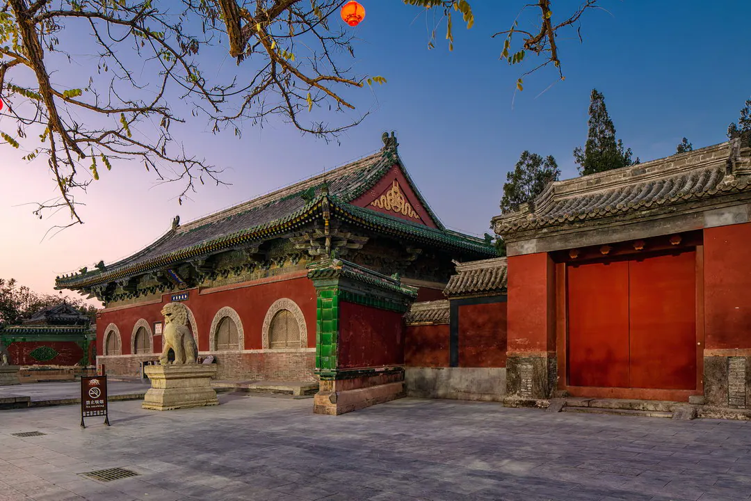 黄昏时分,红墙寺庙大殿有绿琉璃瓦檐,庭院中立有一尊石狮