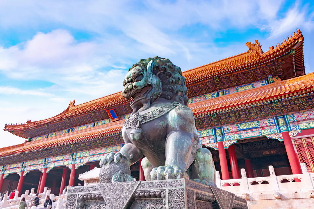 Bronze guardian lion with a paw on a ball, before a red-pillared hall in the Forbidden City