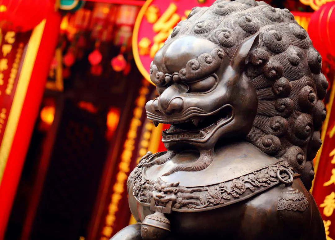 銅獅鬃毛捲曲，頸戴花環，位於紅色寺廟入口之前