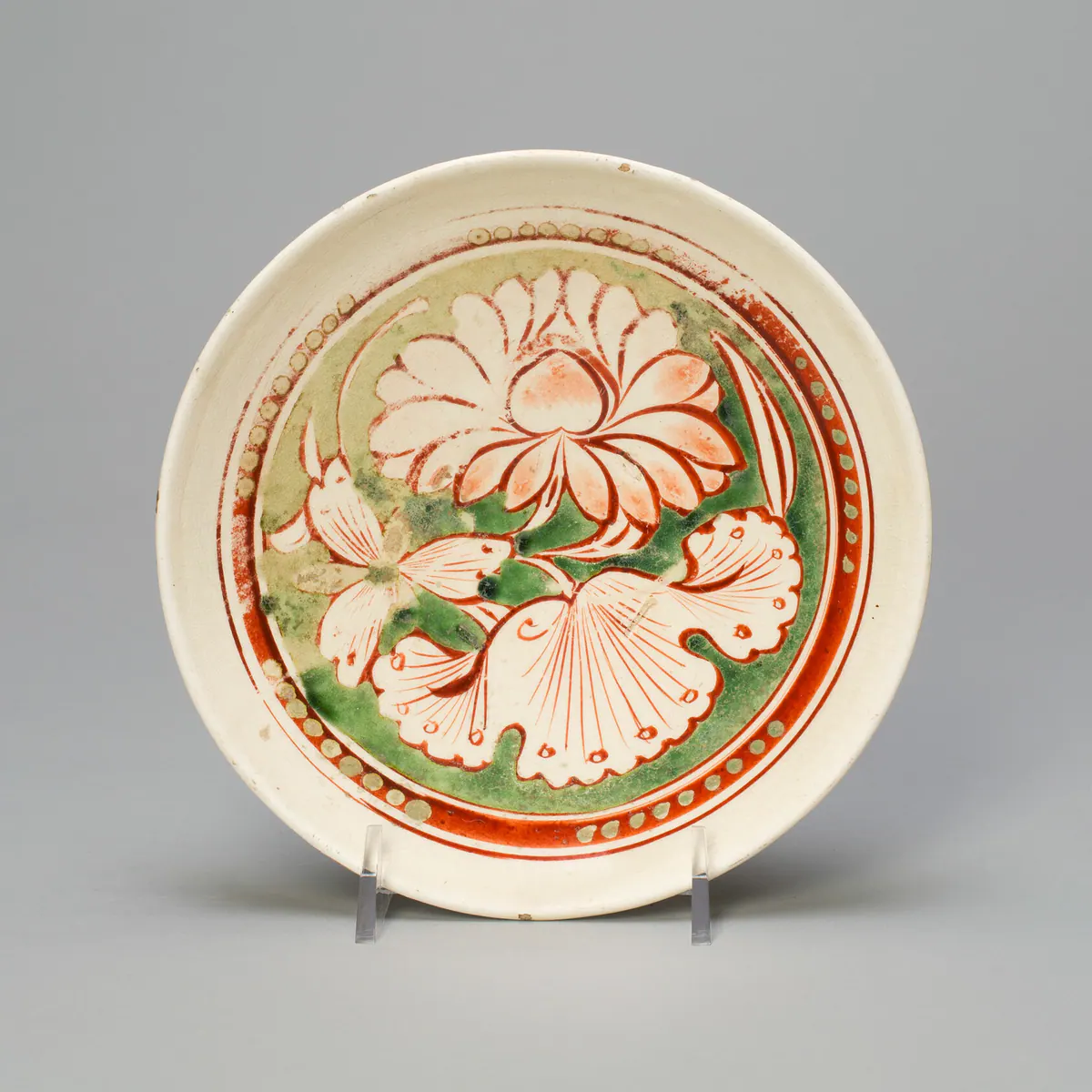 A shallow Cizhou ware bowl, shown upright on a clear display stand, its cream ground painted in red, yellow and green enamels with a large lotus flower and leaves inside a dotted border.