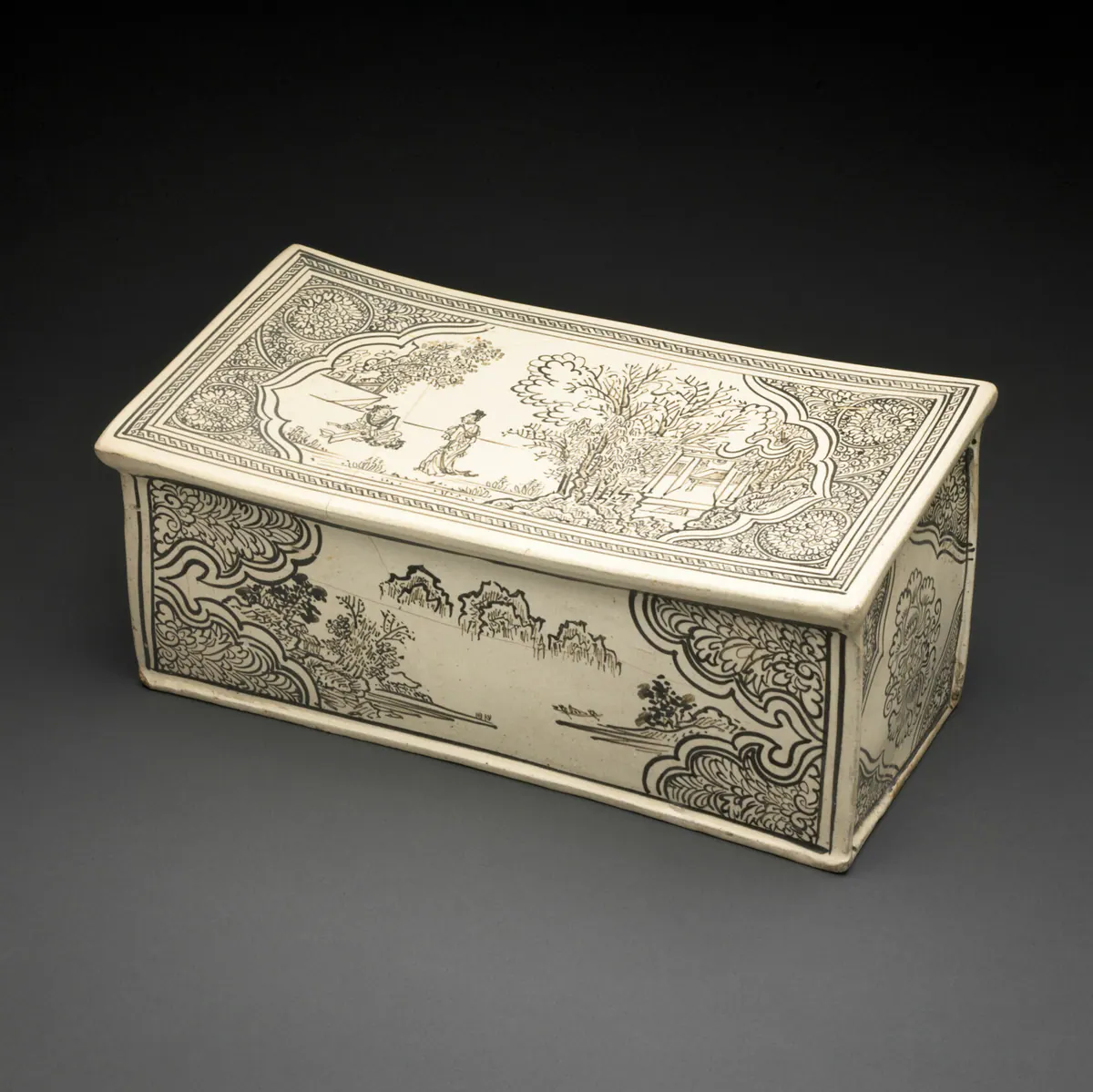 A rectangular Cizhou ware pillow with a cream slip ground, painted in dark brown with two figures in a garden on the top panel and a river landscape on the side, framed by floral borders.
