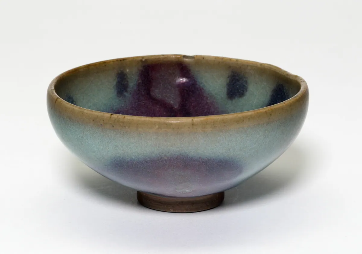 A small, deep Jun ware bowl with a thick, opaque pale blue glaze splashed with purple inside and out, the glaze thinning to olive brown at the rim above a bare foot.