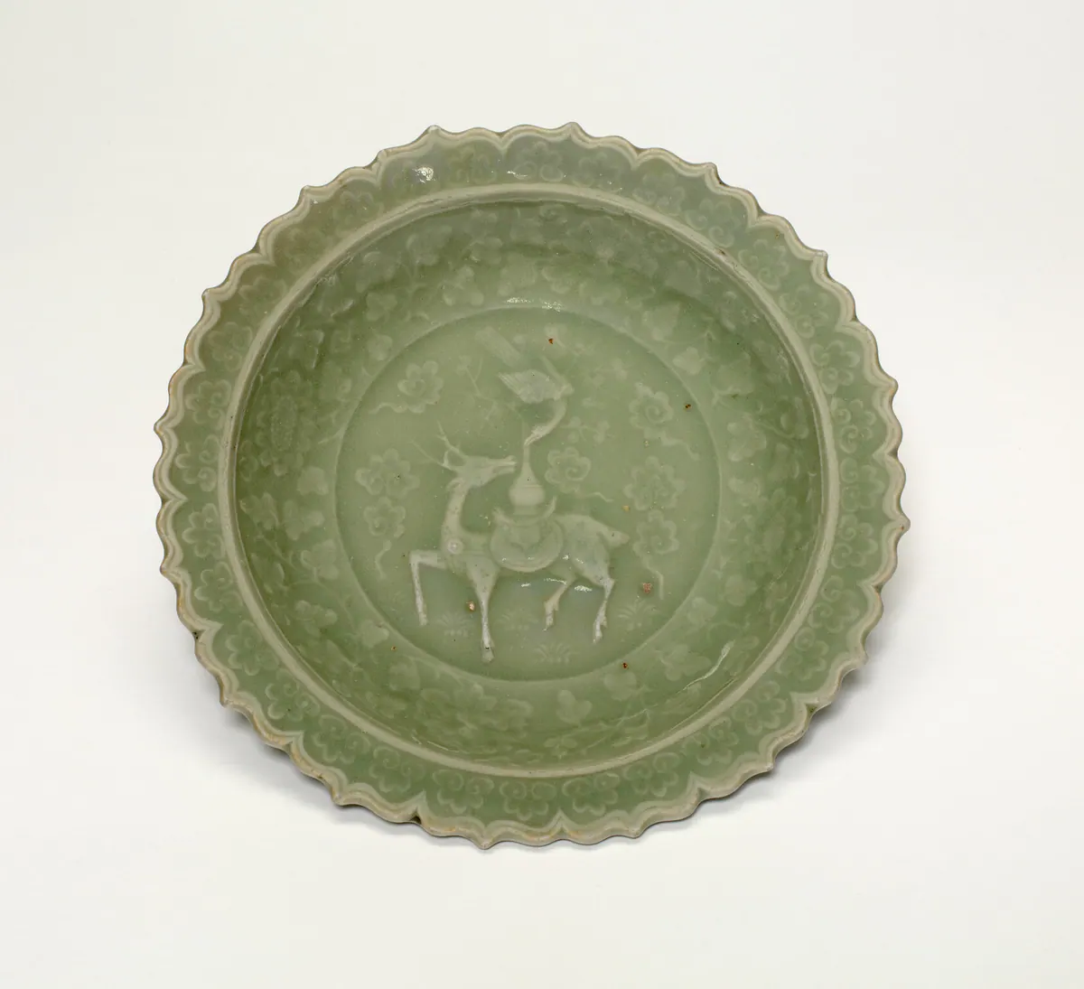 A Longquan celadon dish with a scalloped rim, seen from above, moulded in the centre with a deer and a crane among clouds and flowers, under a grey-green glaze.