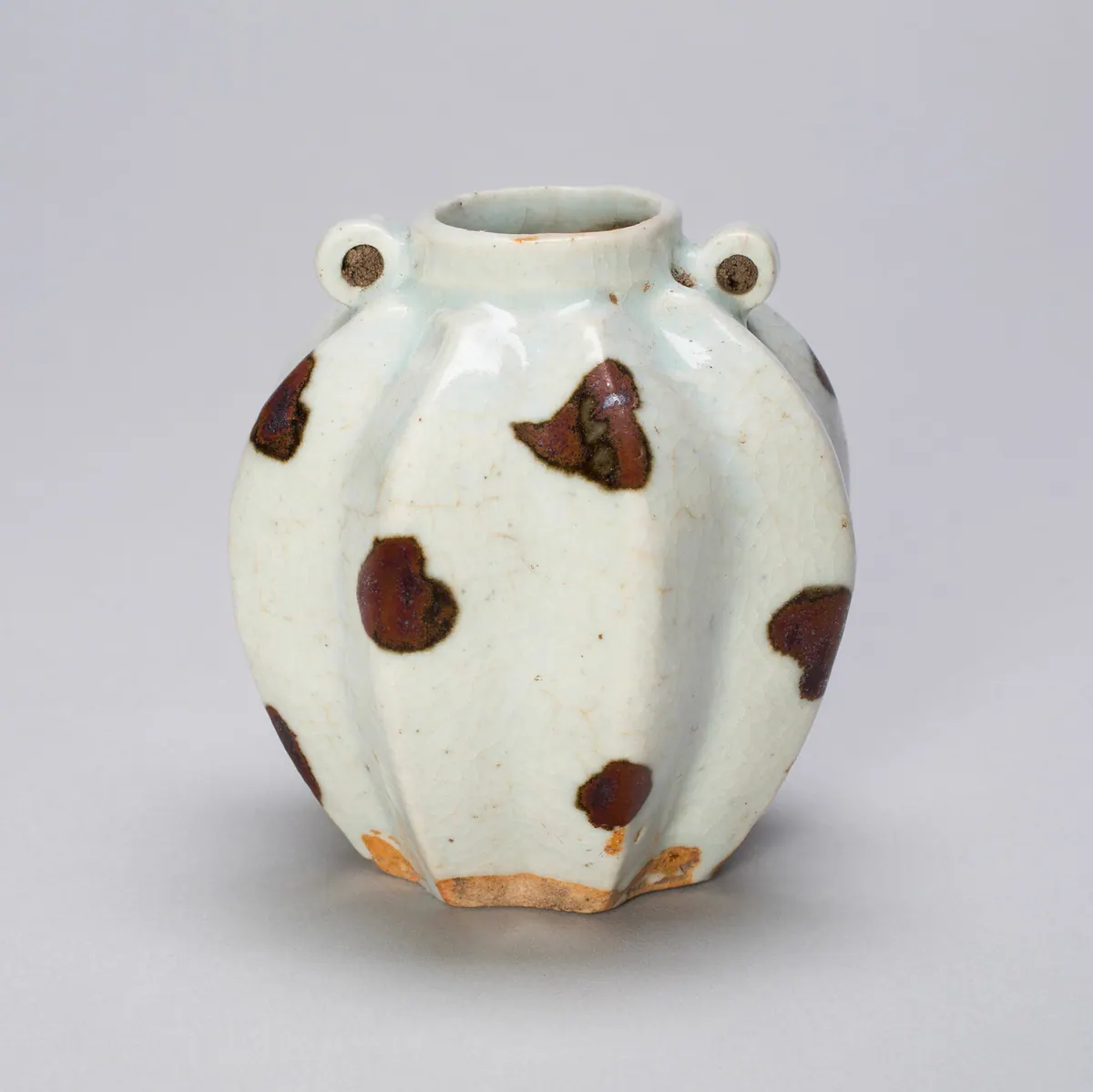 A small lobed qingbai jar with two loop handles at the shoulder, its bluish-white crackled glaze splashed with iron-brown spots, the unglazed base fired orange.