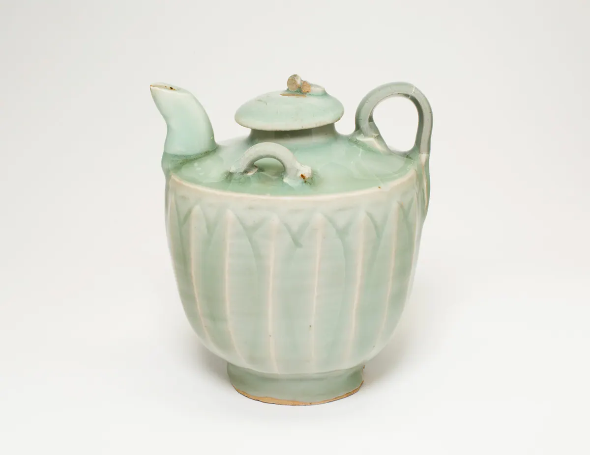 A small celadon ewer with a curved spout, loop handle and domed lid, its body moulded with tall lotus petals under a pale bluish-green glaze.