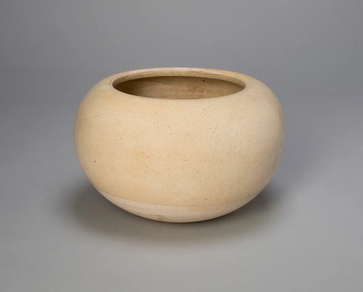 A globular stoneware bowl with an inturned rim, covered in a pale cream glaze that stops short of the base, photographed on a plain grey ground.