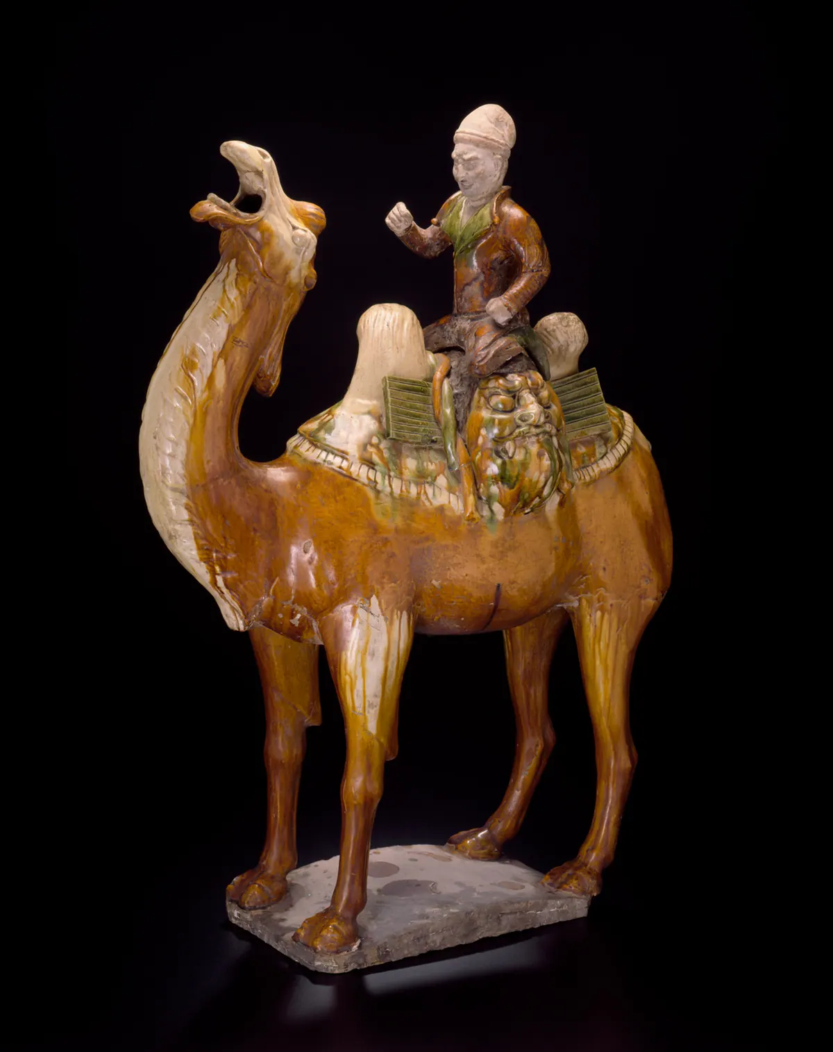 A sancai tomb figure of a Bactrian camel with its head raised and mouth open, glazed amber and cream, carrying a rider in a tall white cap seated on a pack between the humps.