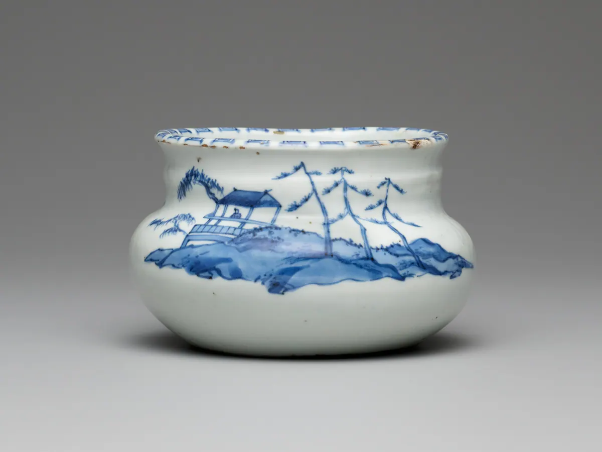 A squat blue and white water jar painted with a pavilion, a seated figure and tall pines on a hillside, with a patterned rim showing small brown spots where the glaze has flaked.