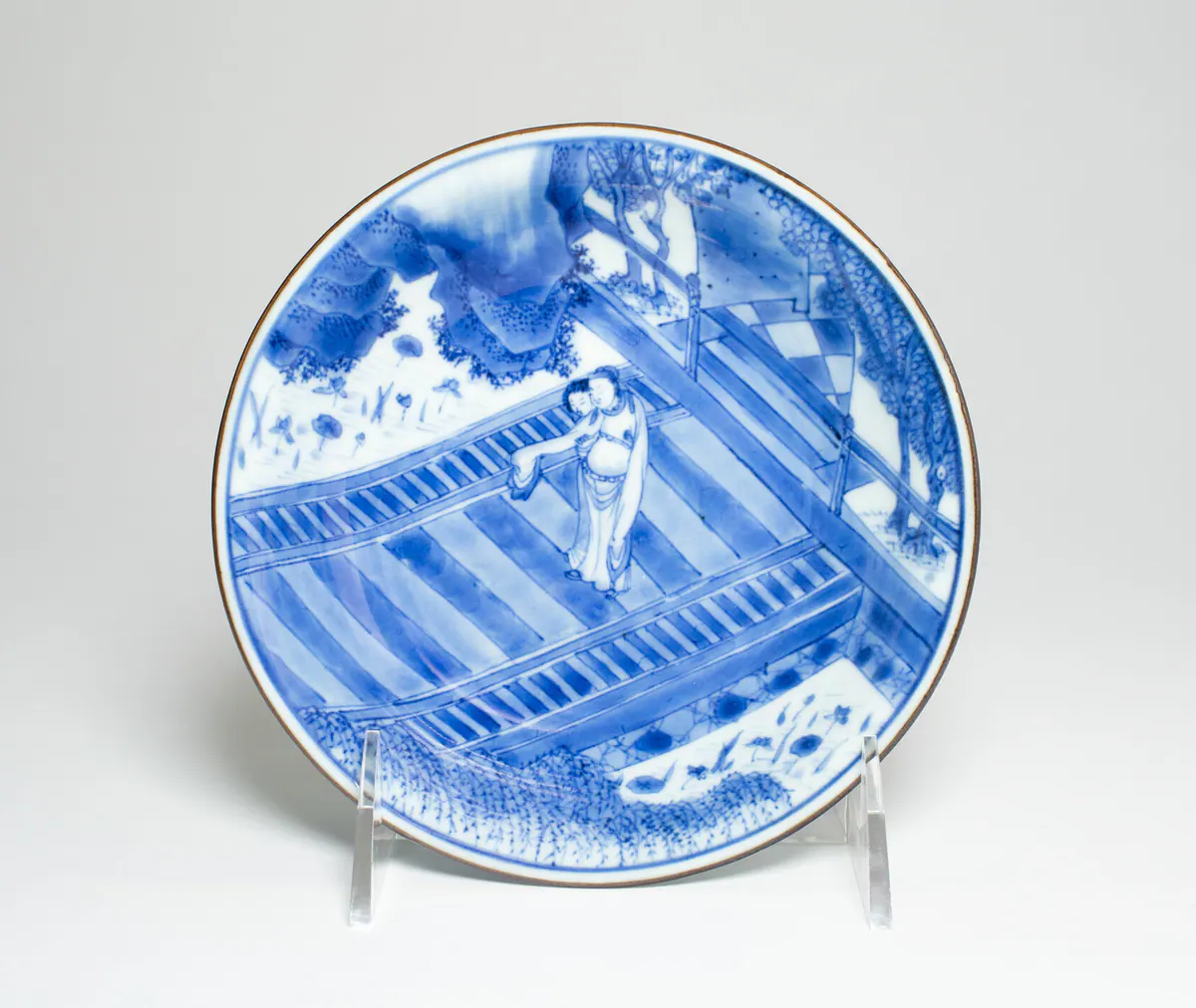 A shallow blue and white bowl seen from above on a stand, painted with two women on a railed terrace beside rocks, trees and a garden pond, under a brown-dressed rim.