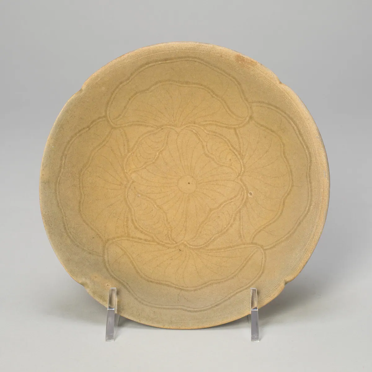 A shallow lobed Yue ware dish seen from above, incised with overlapping lotus leaves around a central flower, under a pale olive-green glaze, on a clear stand.