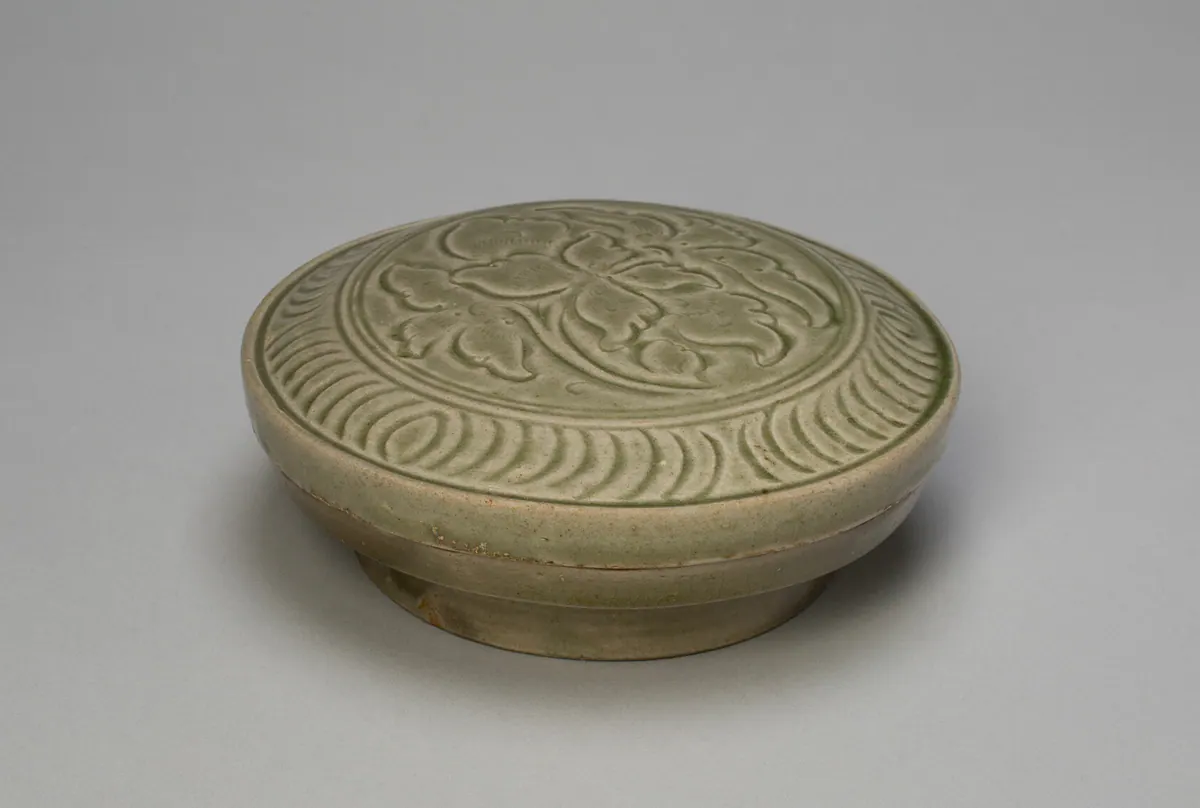 A round Yue ware covered box, the lid carved with a large peony within a border of curved strokes, under an olive-green glaze.