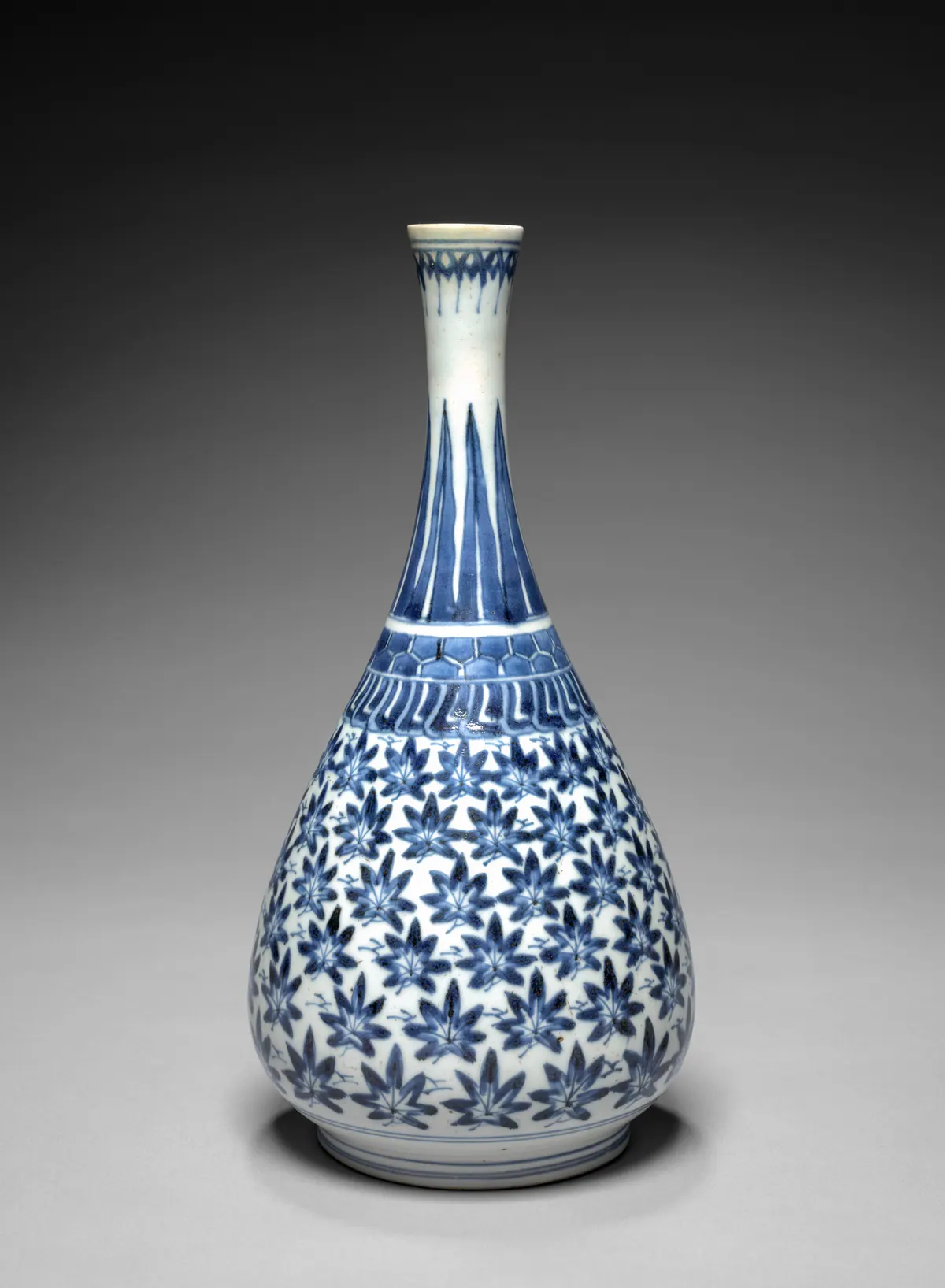 A tall Japanese Arita blue and white bottle with a slender neck, painted in underglaze blue with stiff leaves on the neck, a band of hexagons at the shoulder and maple leaves over the body.
