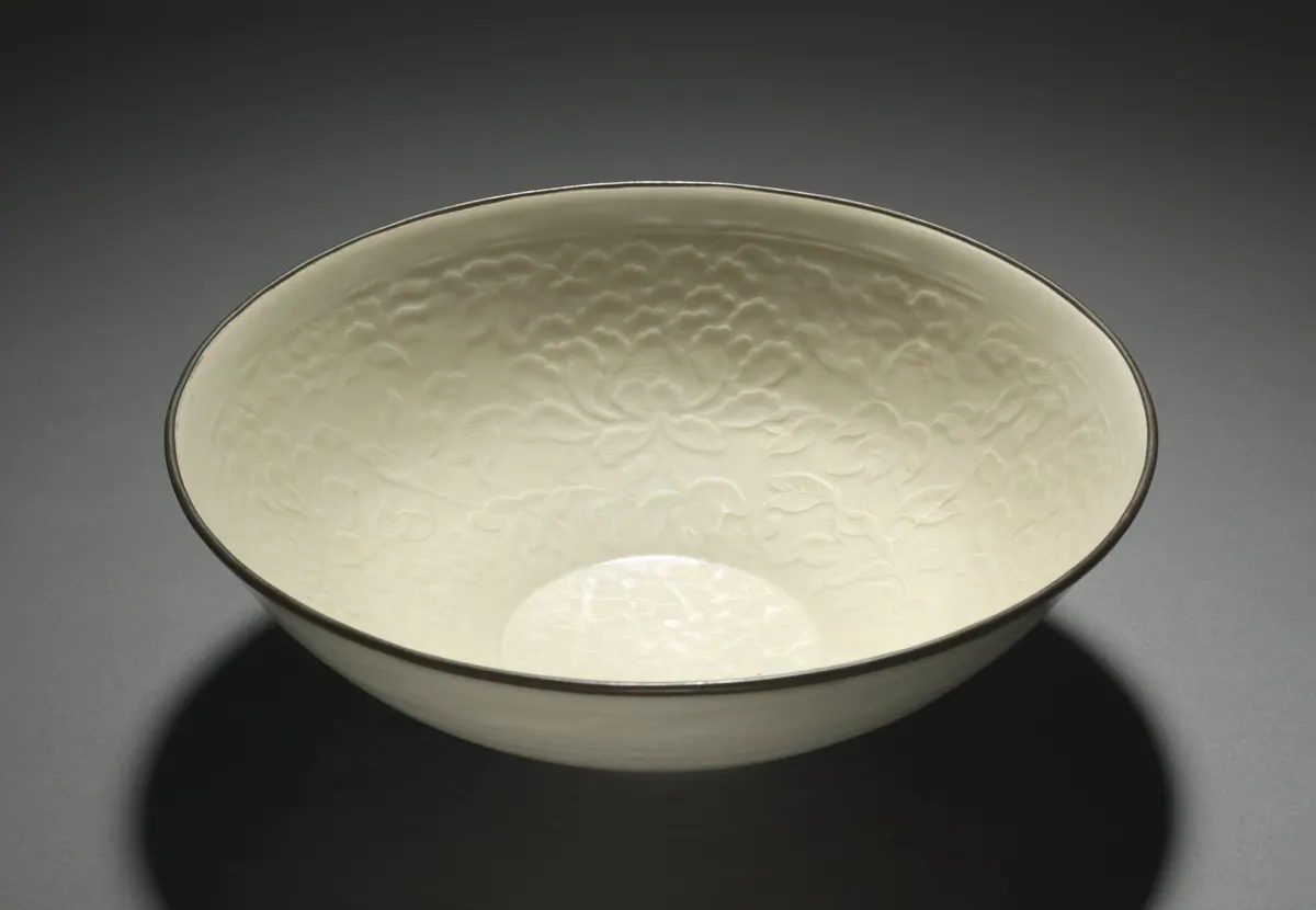 A large Ding ware bowl with a warm ivory glaze, its interior moulded with dense peony scrolls, the bare rim bound with a dark metal band.