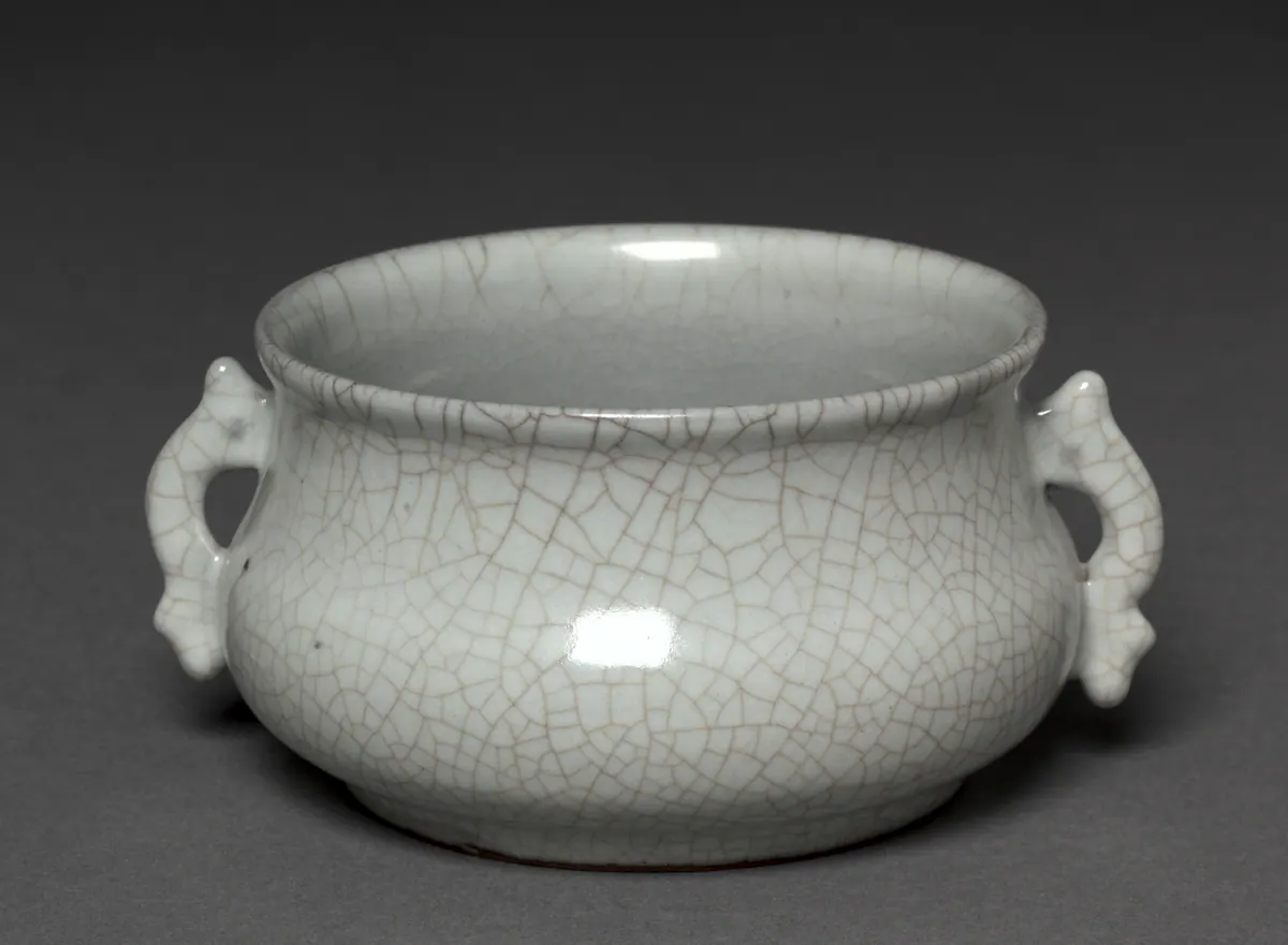 A small Guan ware incense burner of squat round form with two handles, its thick, pale grey glaze covered in a dense network of light brown crackle.