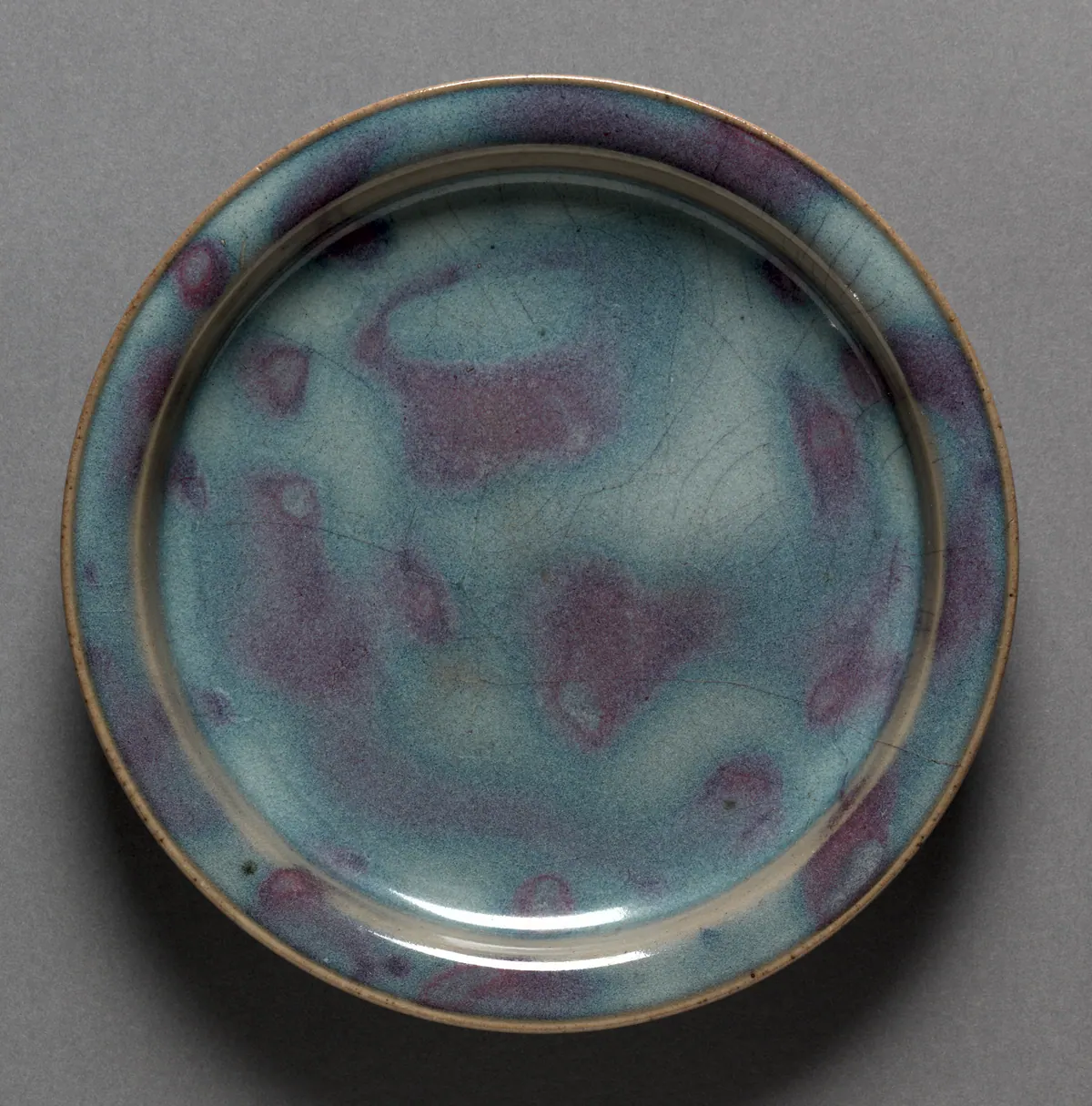 A small Jun ware plate seen from above, its thick opalescent sky-blue glaze broken by soft purple splashes, with a few crackle lines and a buff rim where the glaze thins.