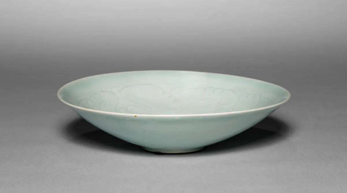 A shallow, wide qingbai dish on a small foot, the inside carved with flowing floral and wave-like scrolls where the pale blue glaze gathers more deeply.