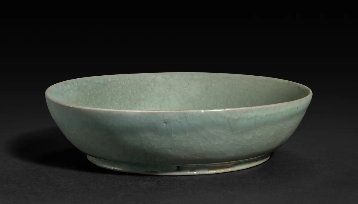 A small, shallow Ru ware brush washer with an opaque bluish-green glaze covered in a fine network of crackle, standing on a low foot against a dark background.