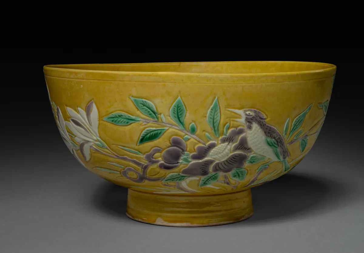 A deep bowl on a tall foot with a mustard-yellow ground, incised and filled in green, aubergine and white with a bird perched among flowering branches and leaves.