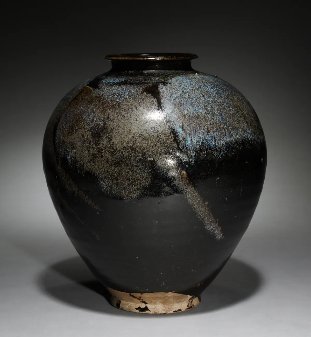 An ovoid stoneware jar under a glossy black glaze, its shoulder streaked with splashes of mottled bluish-white glaze, the foot left unglazed.