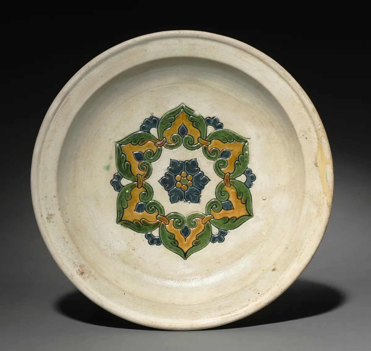 A shallow sancai plate seen from above, its cream ground centred on an impressed six-petalled floral medallion glazed in green, amber and blue.