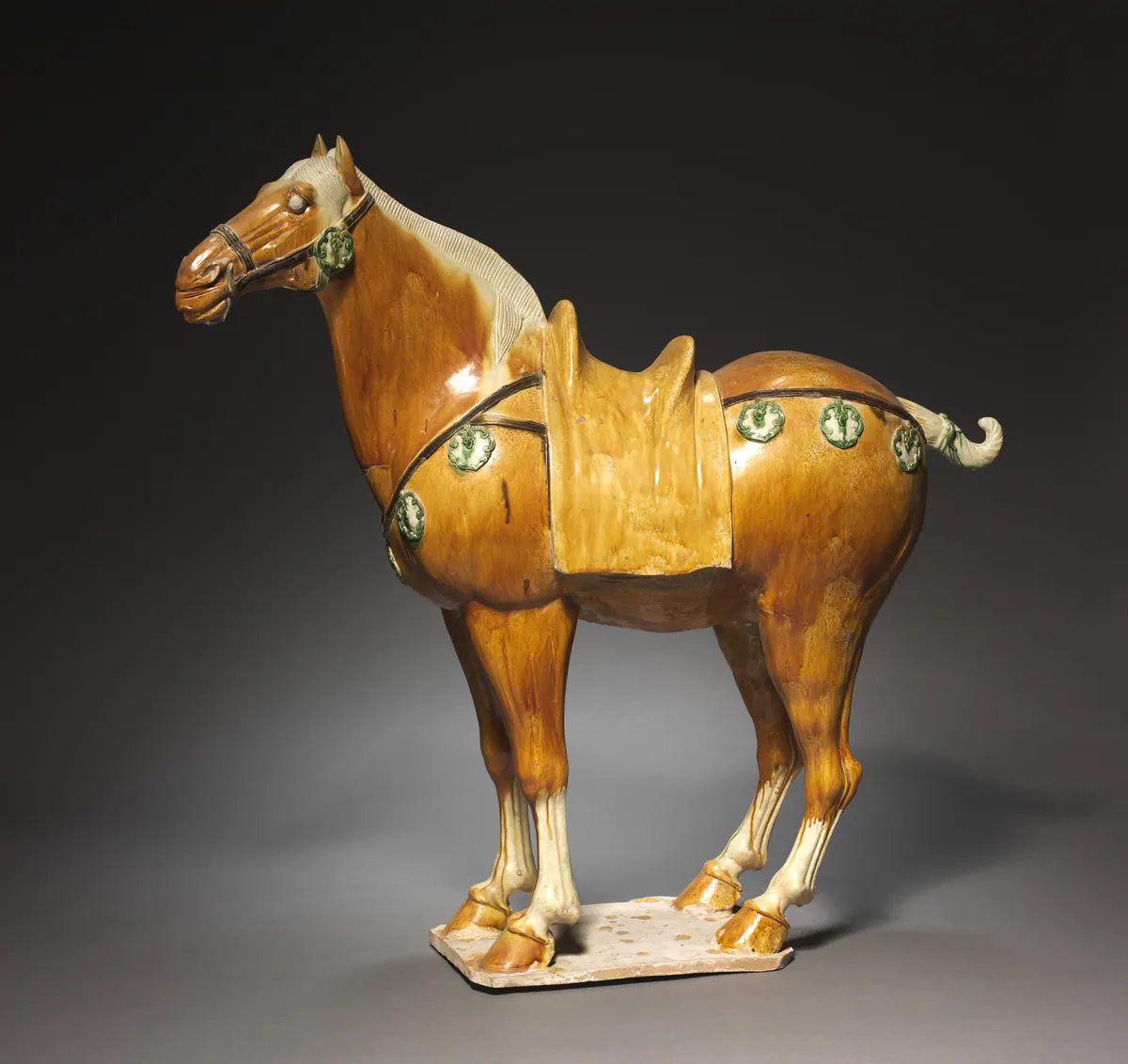 A standing sancai tomb horse glazed in amber with a cream mane and legs, its harness hung with green and cream medallions, on a flat rectangular base.
