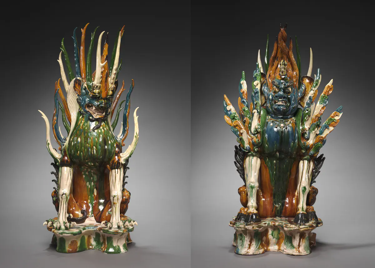 A pair of seated sancai tomb guardian beasts, one with an animal face and one with a human face, both with flame-like crests and wings glazed green, amber, blue and cream.