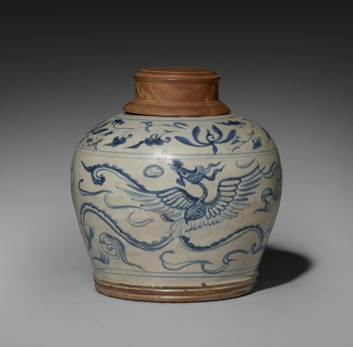 A Vietnamese blue and white jar painted with a phoenix among clouds and a floral band at the shoulder, under a thin, crazed greyish glaze, with a turned wooden cover.