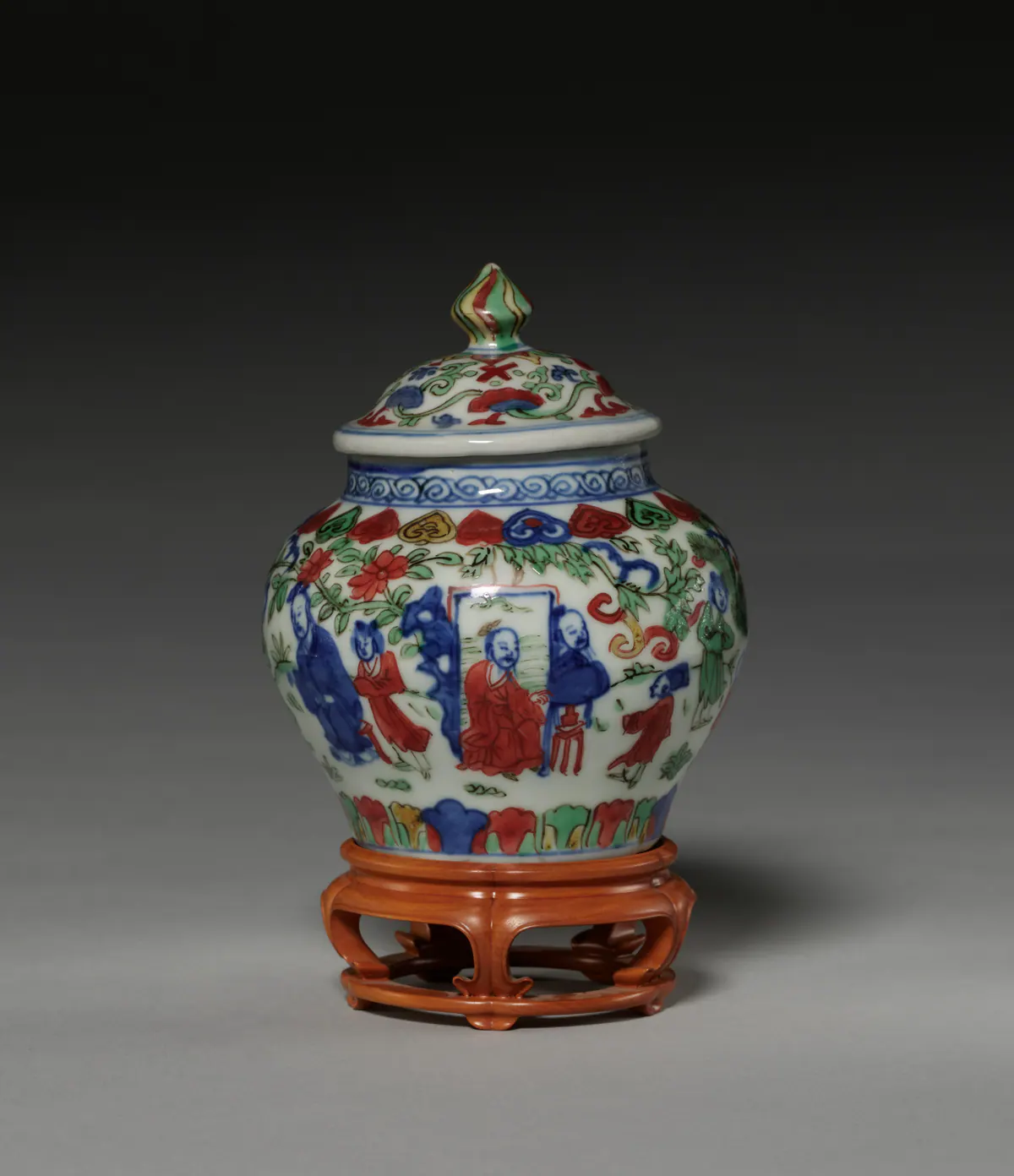 A small Wanli covered jar painted in wucai enamels and underglaze blue with figures in a garden, a band of floral motifs at the shoulder, standing on a carved wooden stand.