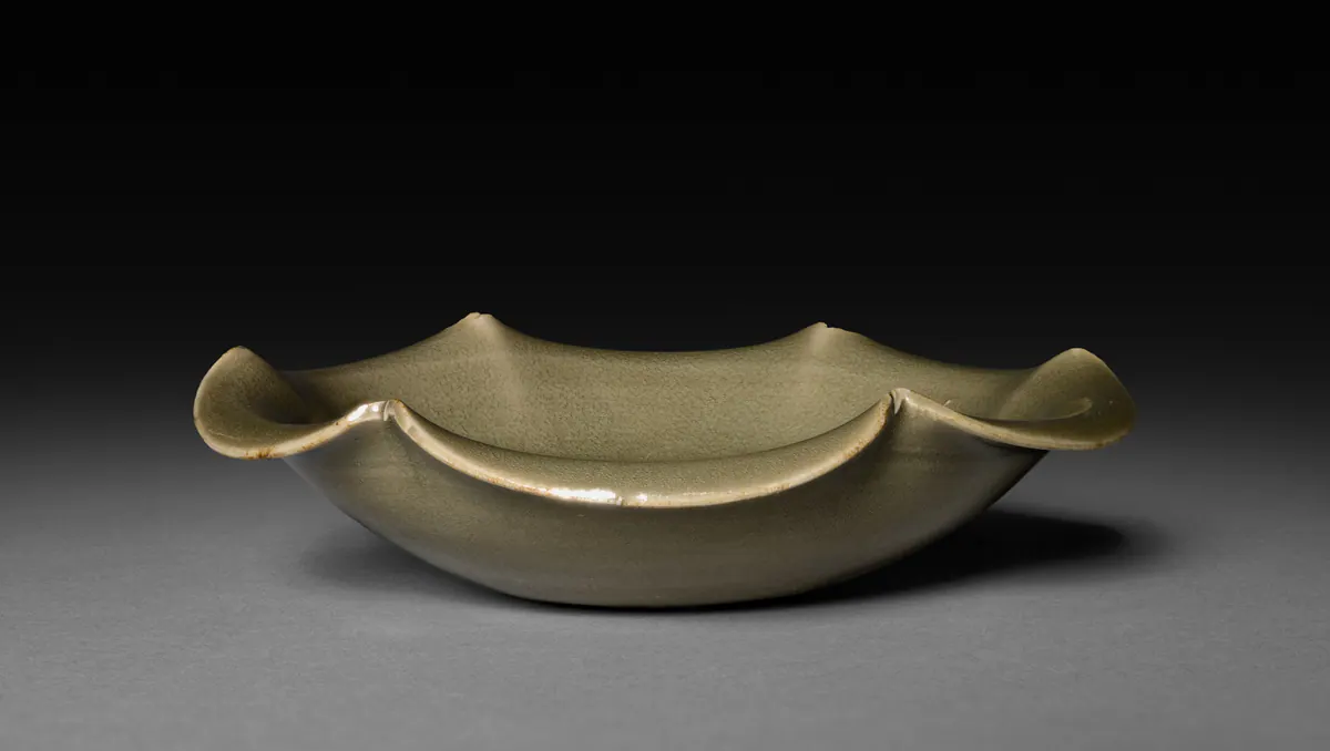 A small Yaozhou celadon saucer seen from the side, with a wide, upturned foliated rim under an even olive-green glaze.