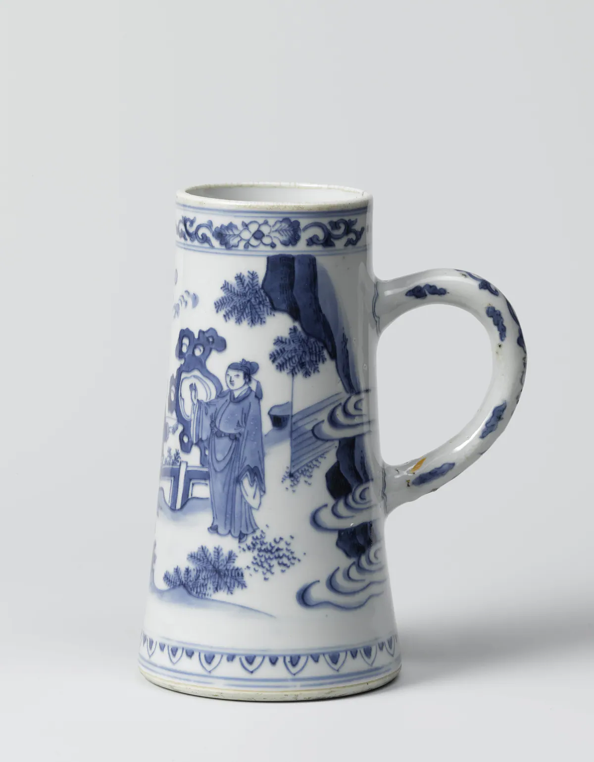 A Transitional blue and white tankard of tapering European form with a loop handle, painted with a scholar by a garden rock and fence, with plants, clouds and a tall cliff, below a band of flower scrolls.